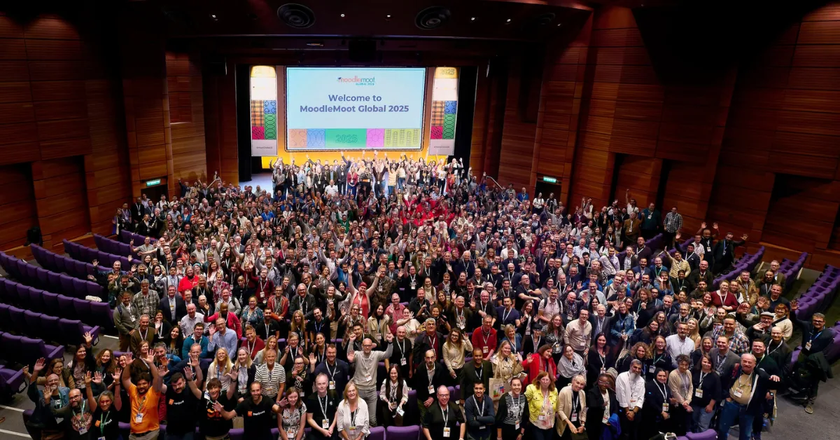 Hundreds of MoodleMoot Global 2025 attendees fill a lecture theatre, many waving at the camera, in front of the event's welcome slide.