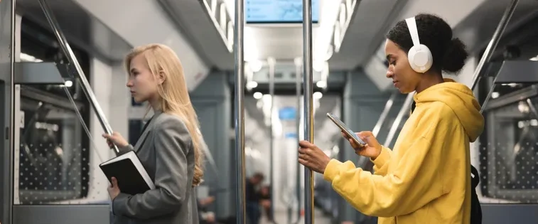 Two women in a subway, one reading and the other listening to music on headphones.