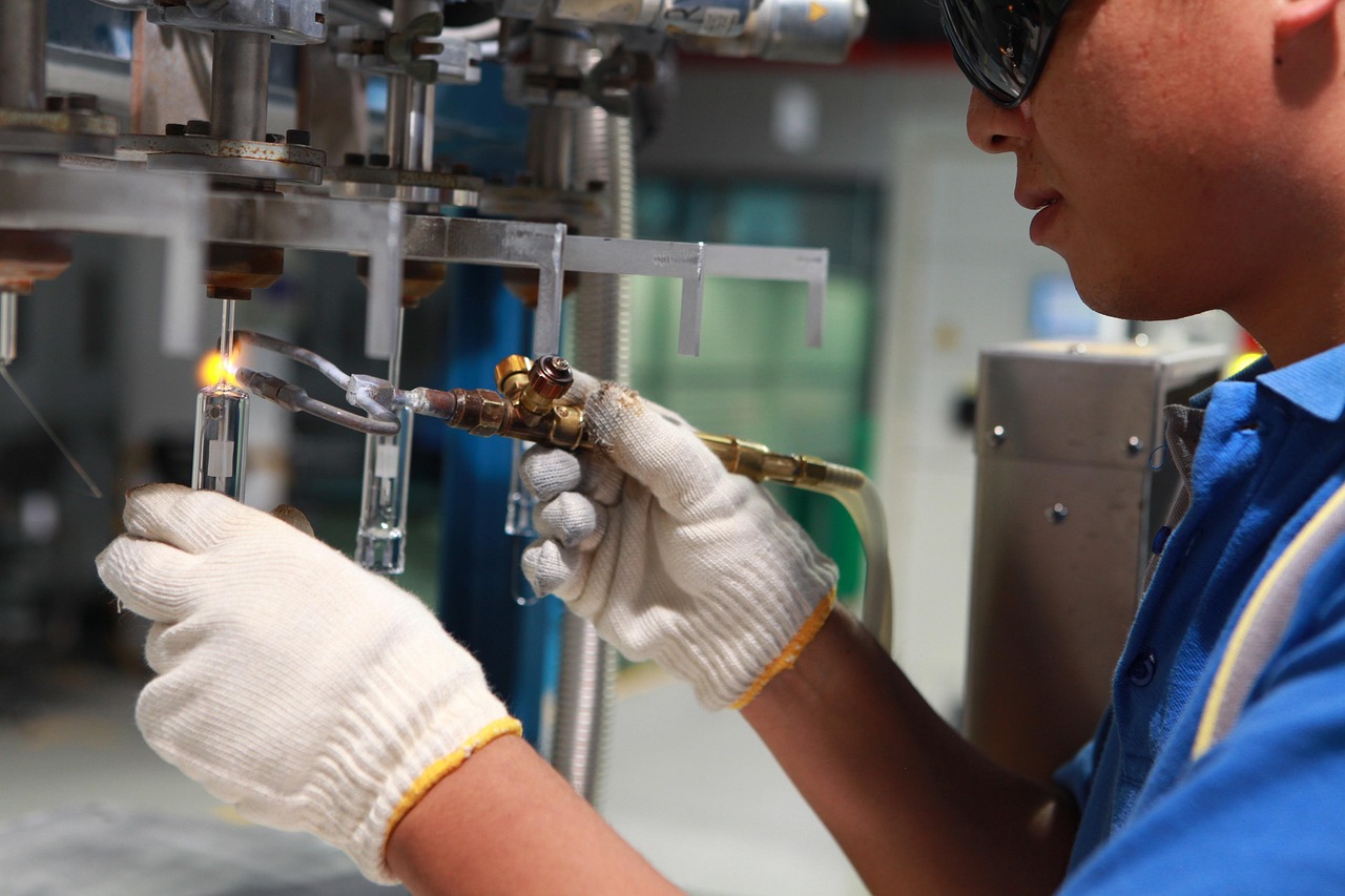 Lavoratore che indossa guanti e occhiali di sicurezza mentre usa una torcia a gas per saldare componenti in vetro in un ambiente industriale.
