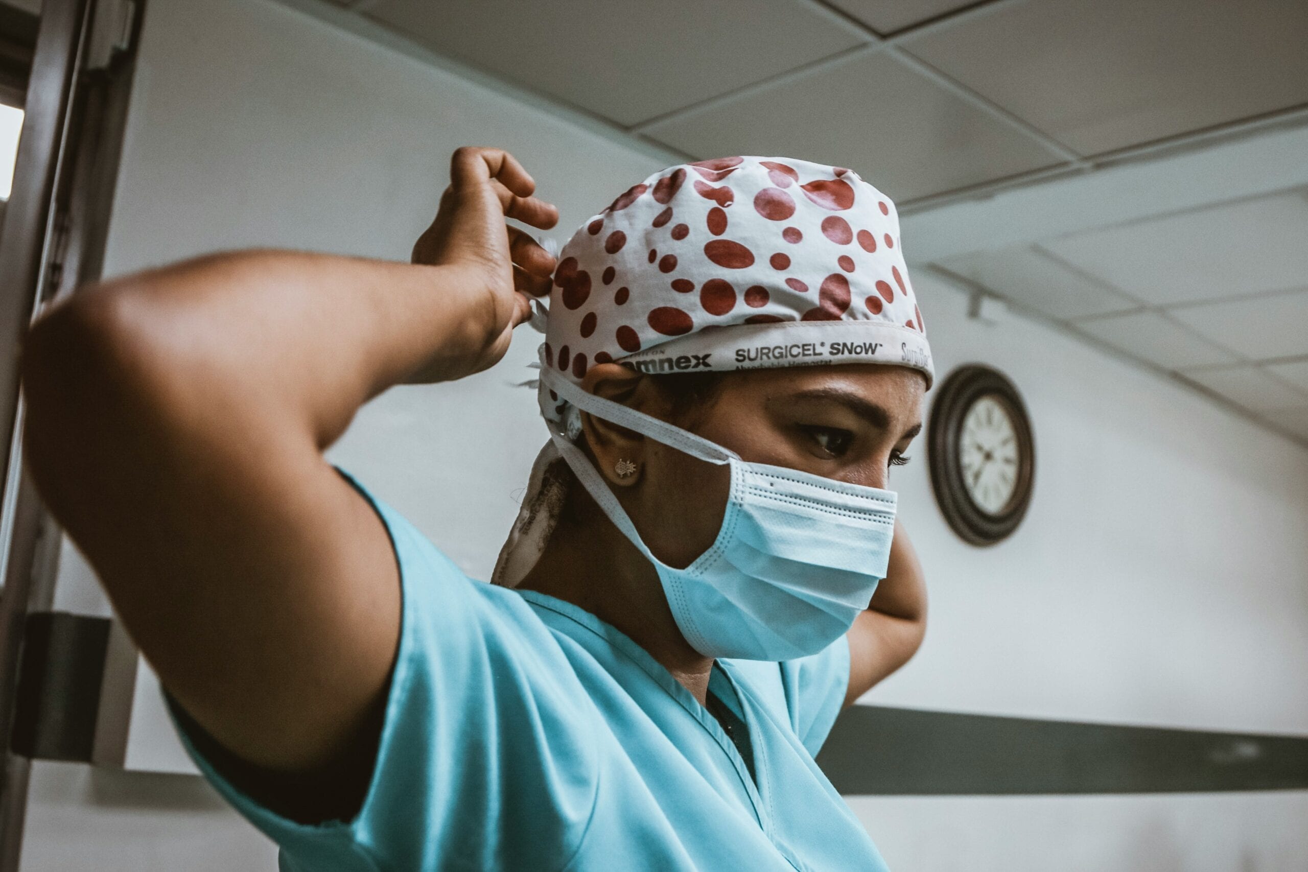 Woman wearing blue surgical scrubs and cap, tying a white face mask behind her head.