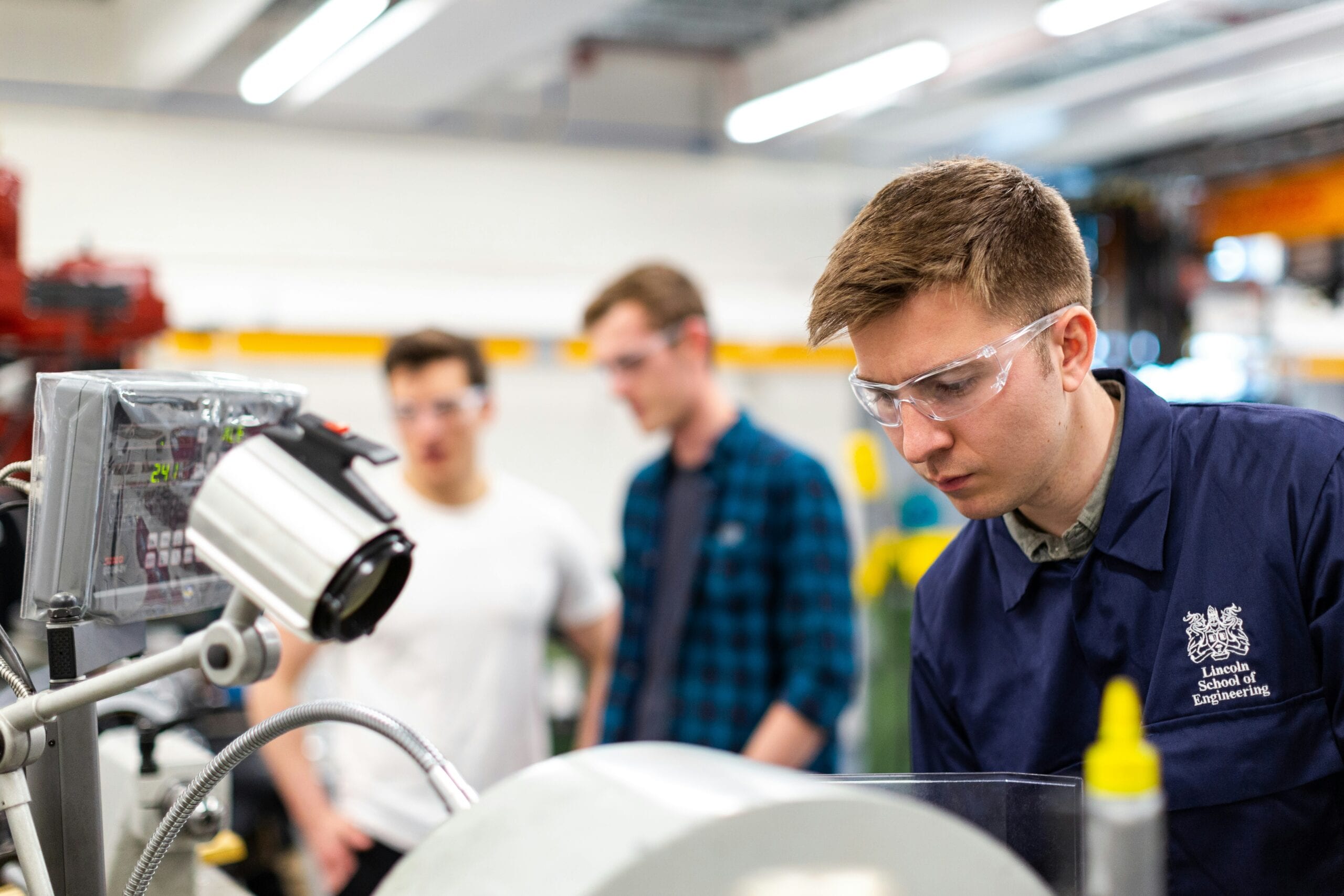 Étudiants en ingénierie, portant des lunettes de protection, debout à côté de machines, représentant l'accessibilité dans la formation professionnelle.