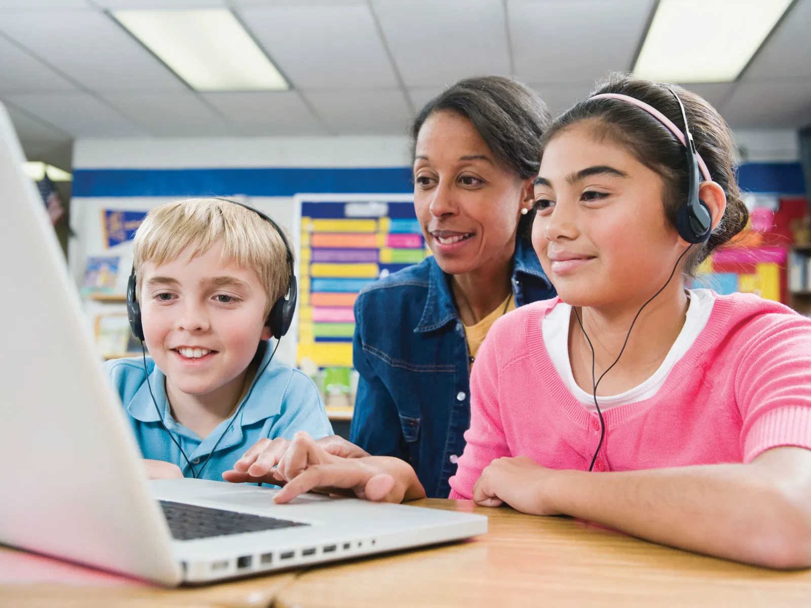 Dois alunos na escola usam fones de ouvido e tecnologia de conversão de texto em fala enquanto fazem seus trabalhos escolares em um computador portátil. O professor está sentado atrás deles, observando como trabalham.