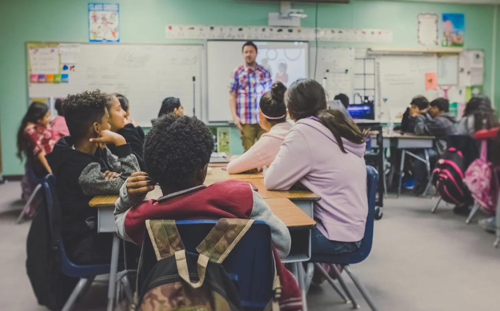 Os alunos estão em uma sala de aula e ouvindo o professor no quadro.
