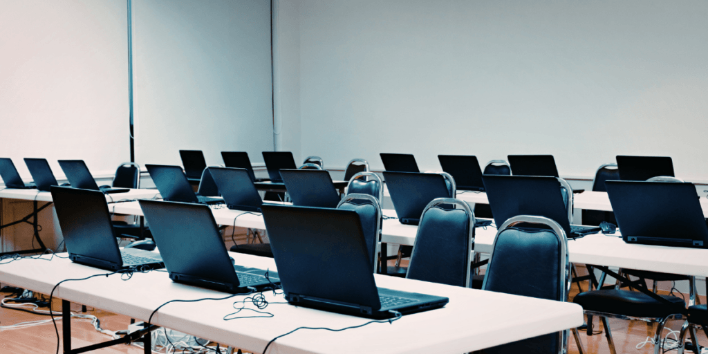 Image d’une salle de classe avec plusieurs ordinateurs portables alignés sur trois rangées de bureaux, représentant des évaluations numériques.
