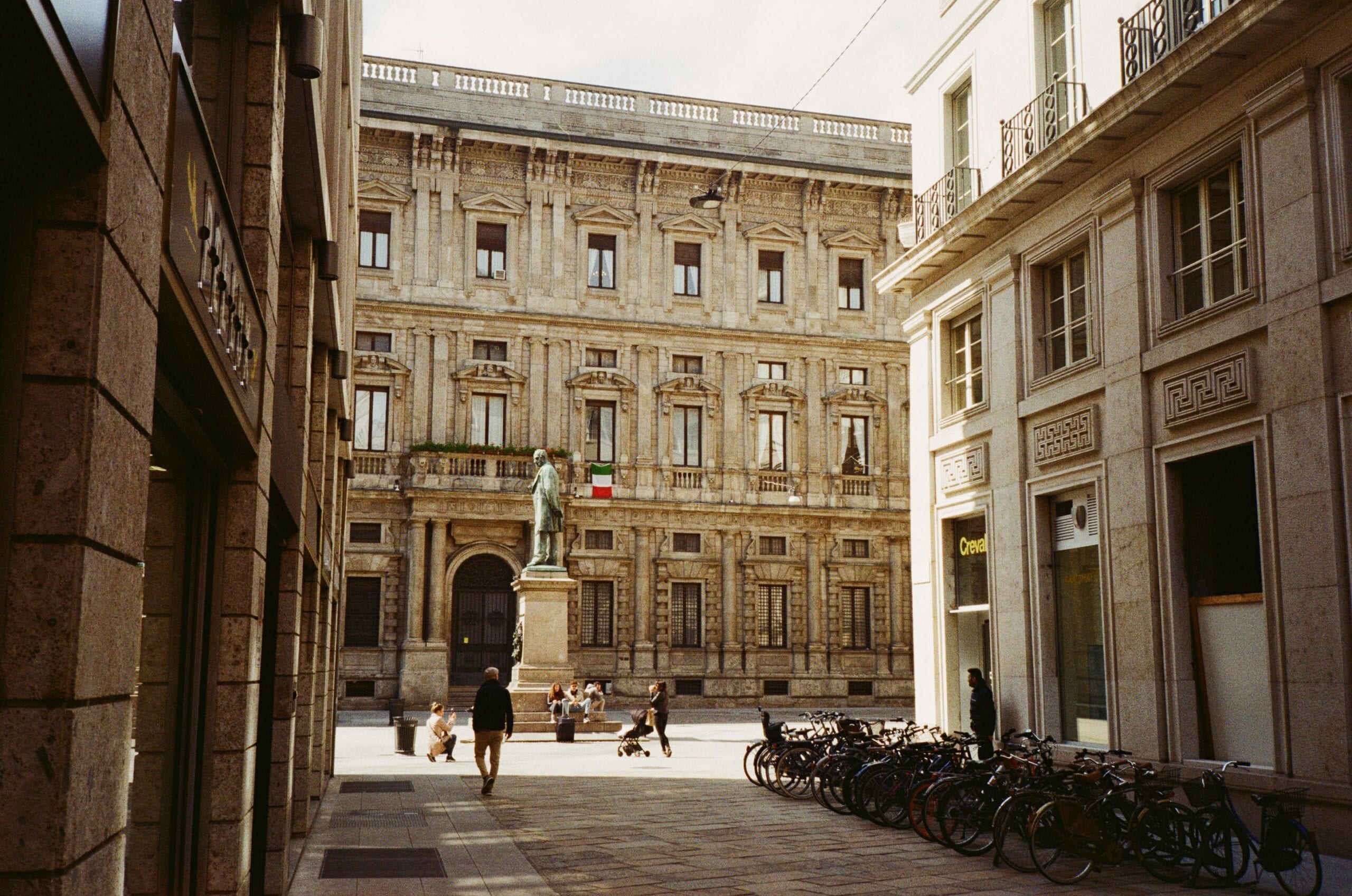 Una via stretta di Milano che si apre su una piccola piazza, dove una statua di bronzo su un basamento di pietra si trova davanti a un palazzo signorile con la bandiera italiana. Alcuni passanti attraversano la pavimentazione e una lunga fila di biciclette e parcheggiata sul lato destro.