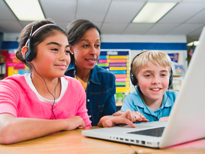 Duas crianças em idade escolar usando fones de ouvido e utilizando texto-para-fala enquanto fazem trabalhos escolares em um laptop. A professora está sentada atrás delas para ver como estão progredindo.