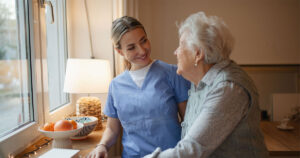 A skilled carer, standing next to, and supporting, an older lady in a nursing home setting.